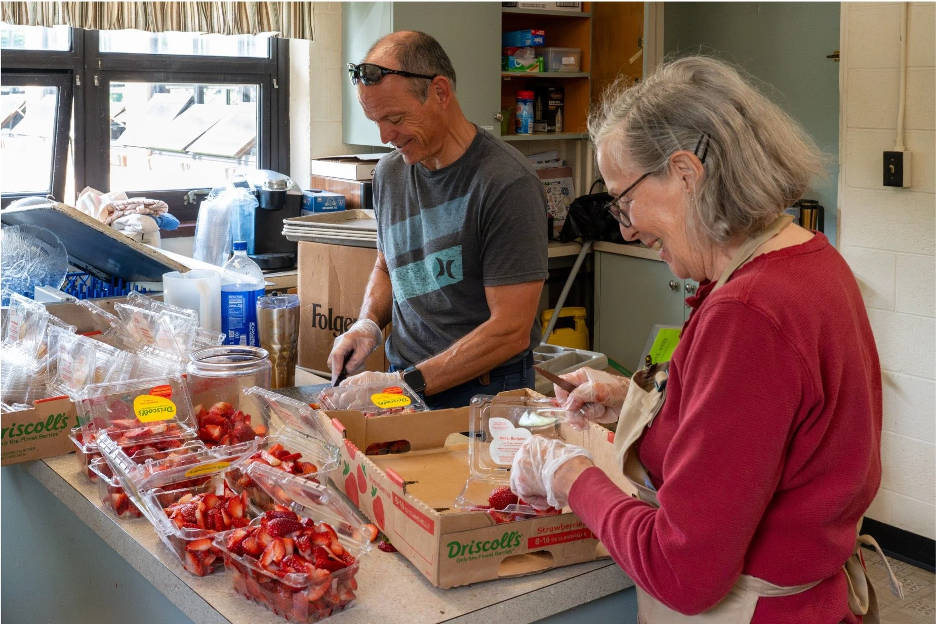 Two volunteers wearing gloves and aprons pack sliced strawberries into clear containers in a commercial kitchen.