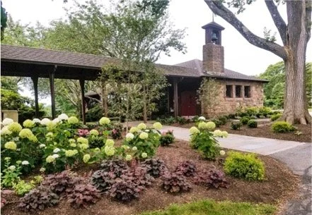 Landscaped garden with hydrangeas and ornamental shrubs beside a rustic stone building with a covered walkway and chimney