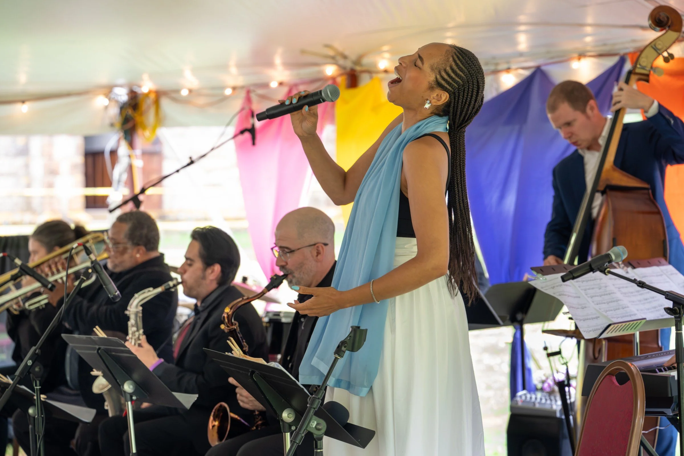 Singer performing with a microphone alongside an orchestra, including a double bass player, under a colorful tent.