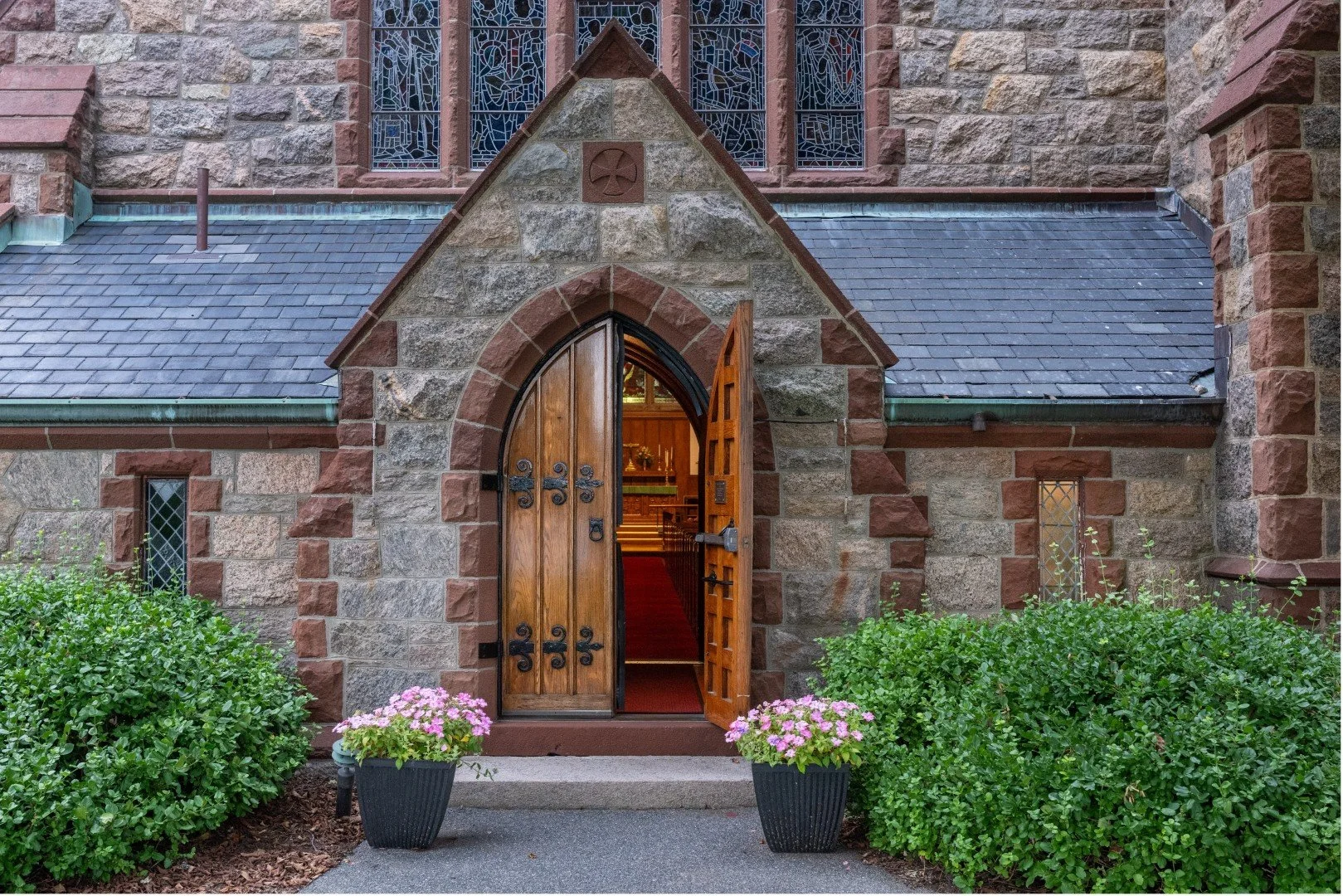 Stone church entrance with open wooden doors, stained-glass windows, manicured hedges, and pink flower planters