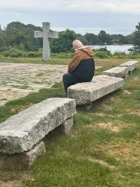 Elderly person sitting on a stone bench near a cross and lake