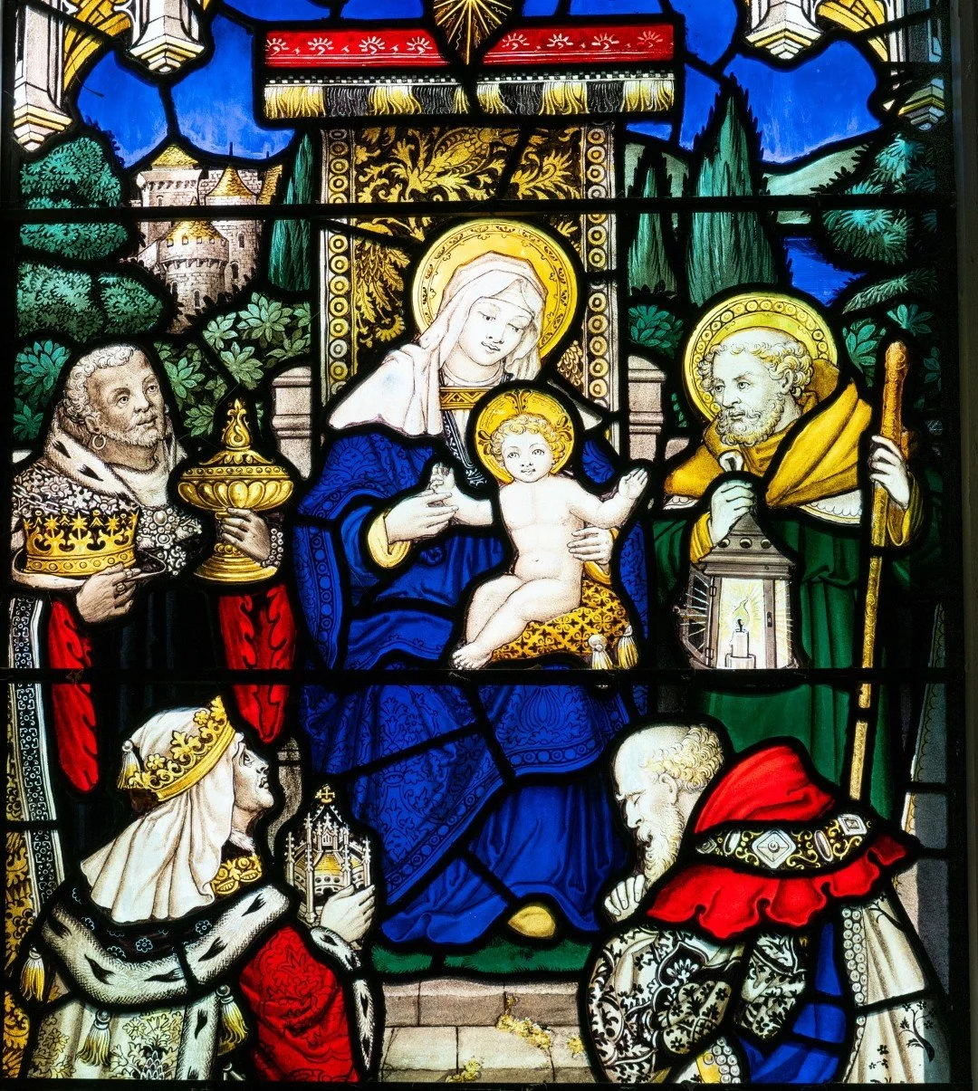 St. B stained-glass window depicting the Virgin Mary holding baby Jesus, surrounded by saints and kneeling worshippers in a medieval castle setting
