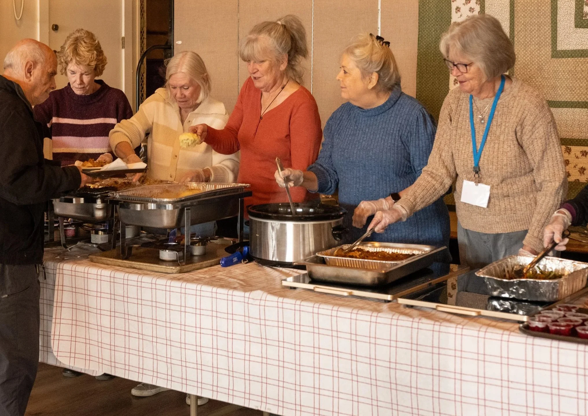 Older adults serving food from chafing dishes and slow cookers at a community buffet.
