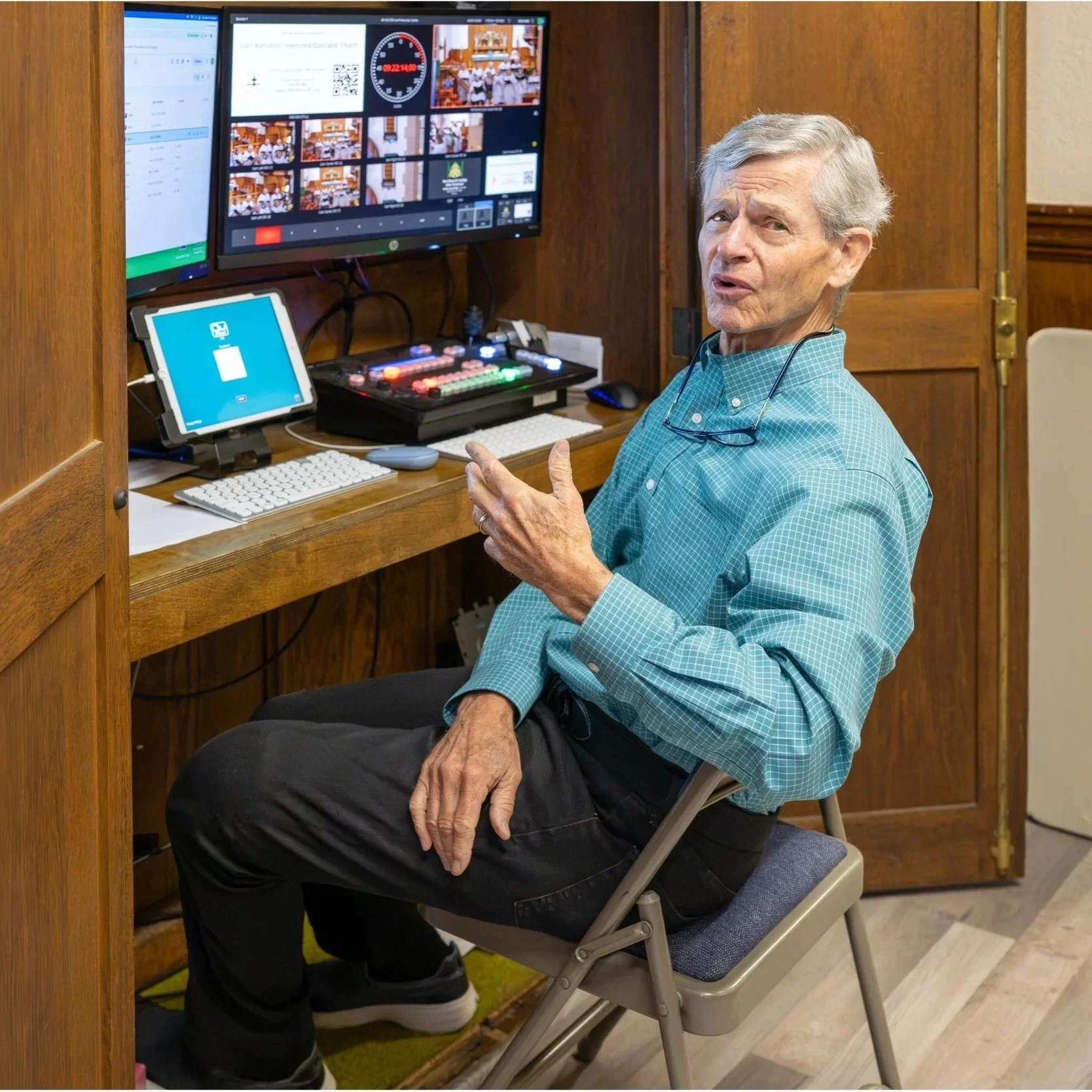 Older man seated beside a video production control desk with monitors, tablet, keyboard, and illuminated switcher.