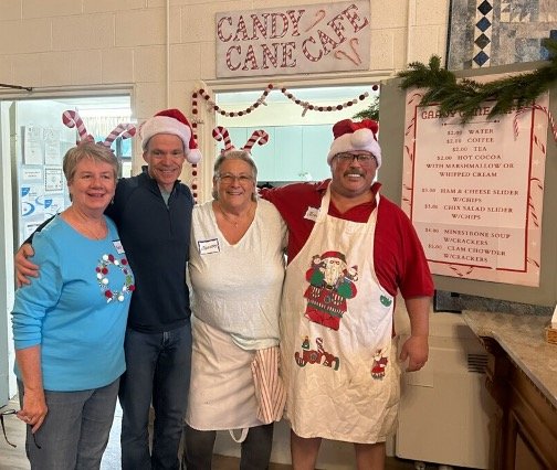 Four people wearing Christmas-themed clothing and Santa hats pose in a decorated community kitchen beside a candy cane café menu.