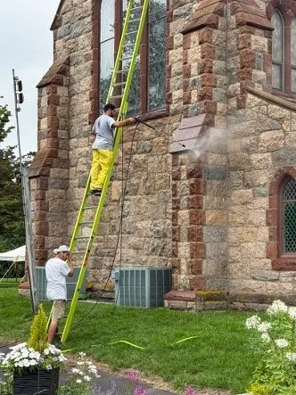 Two workers use a tall ladder to clean a stained-glass church window on a stone church exterior.