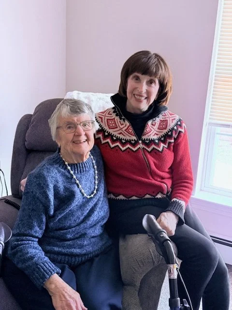 Two smiling women, one older and one middle-aged, seated together indoors; the older woman wears glasses and a blue sweater, while the other wears a red patterned cardigan and holds a cane.