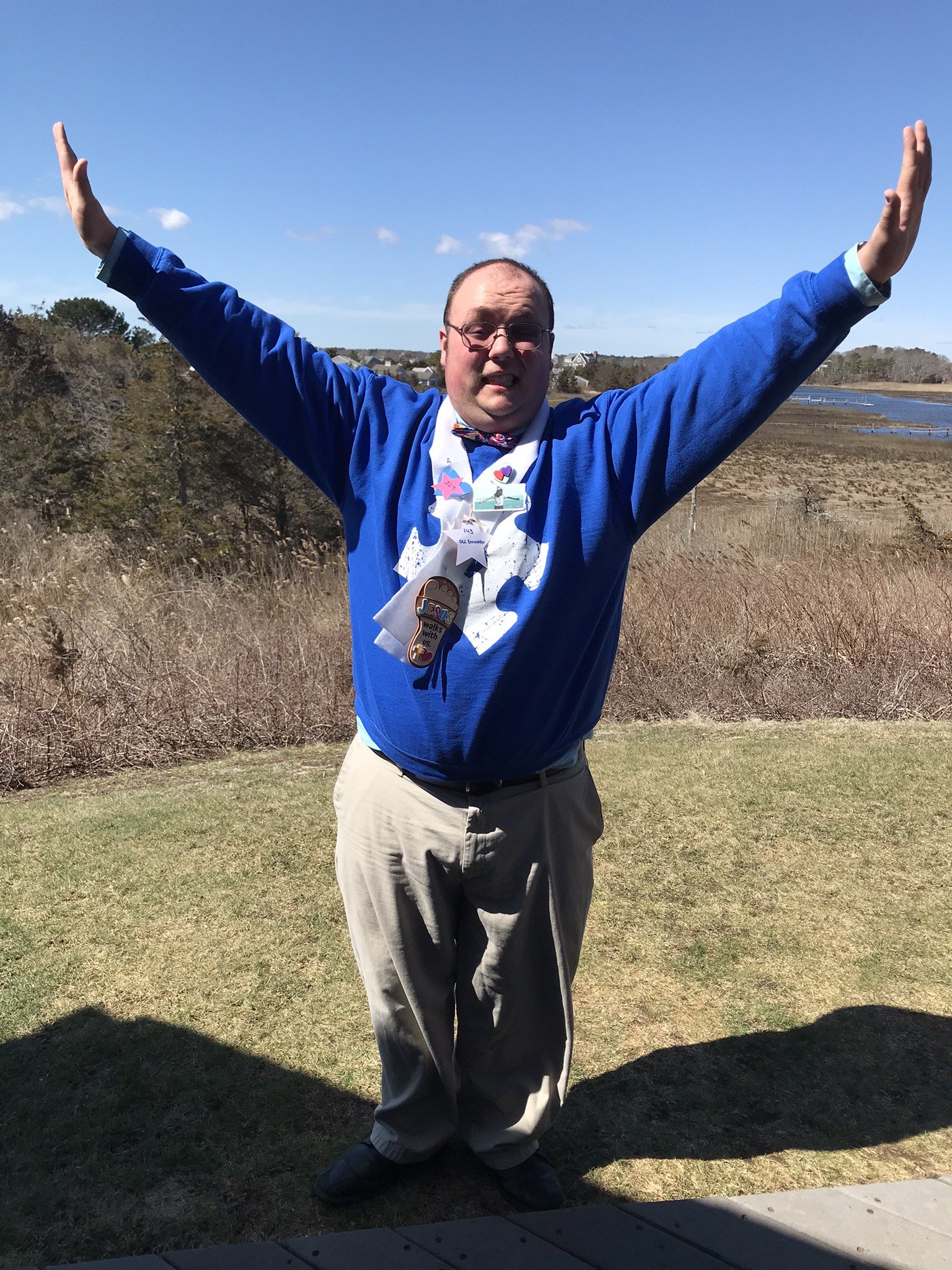 Man wearing a blue sweater, bow tie, and pins stands outdoors with arms raised near a marsh and waterfront.