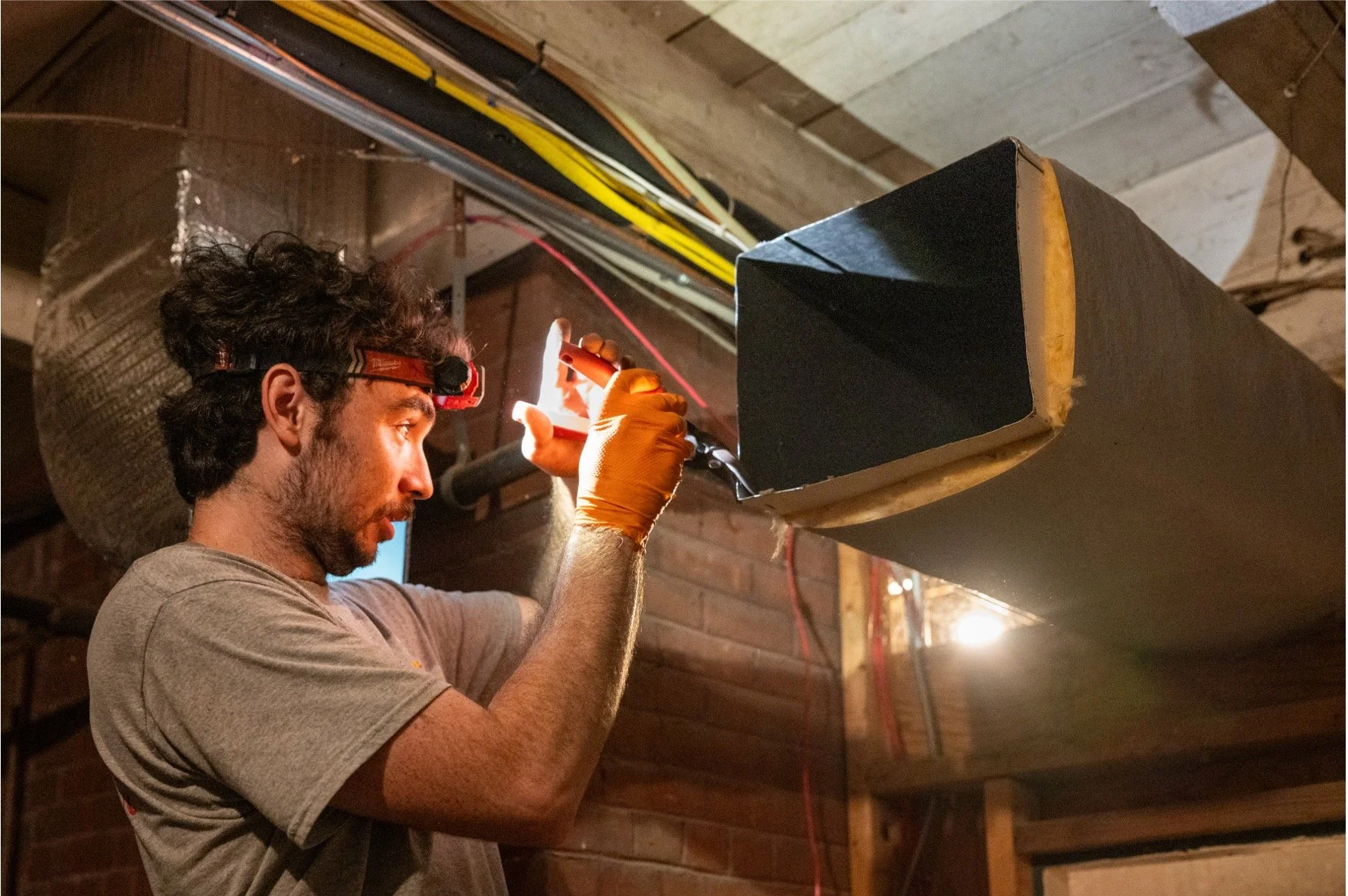 Electrician wearing a headlamp and protective gloves installing ductwork in an unfinished basement