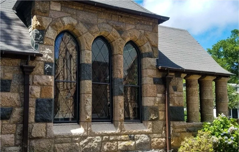 Close-up of our facade with three arched windows and a small porch with four stone columns, part of a house with a gray sloped roof, set in a lush green environment.