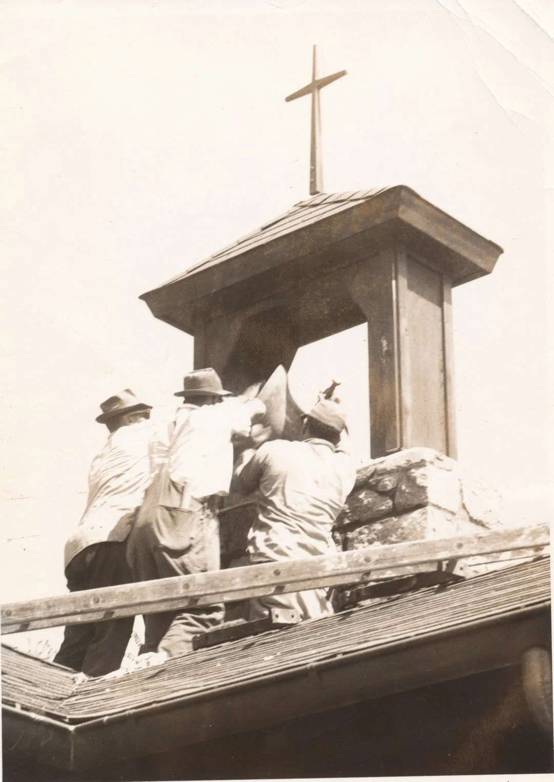 Historic black-and-white photo of three men in hats repairing a chapel roof beneath a large cross.