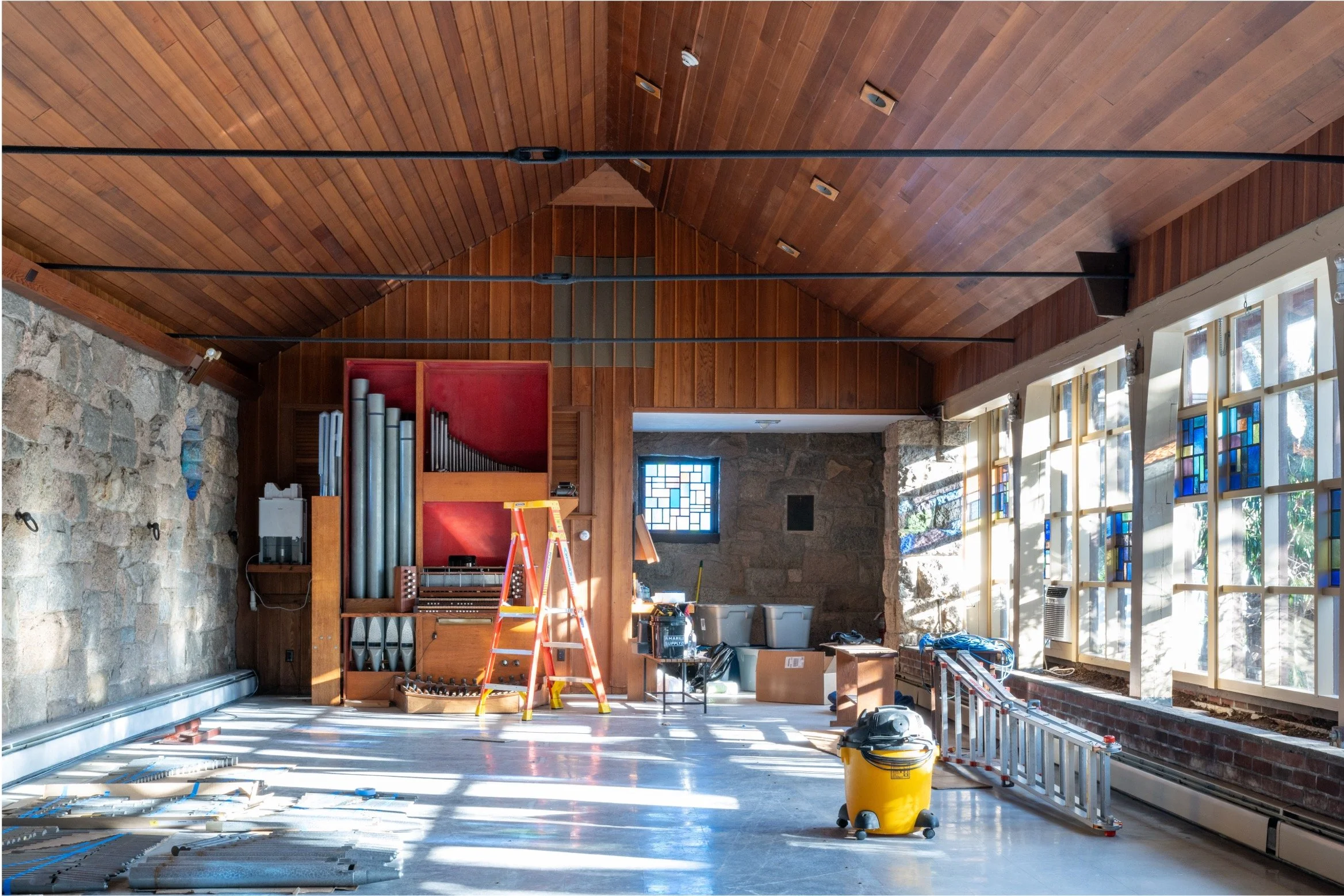 Historic stone building interior under renovation, featuring a pipe organ, wood-paneled vaulted ceiling, stained-glass windows, ladder, and construction equipment