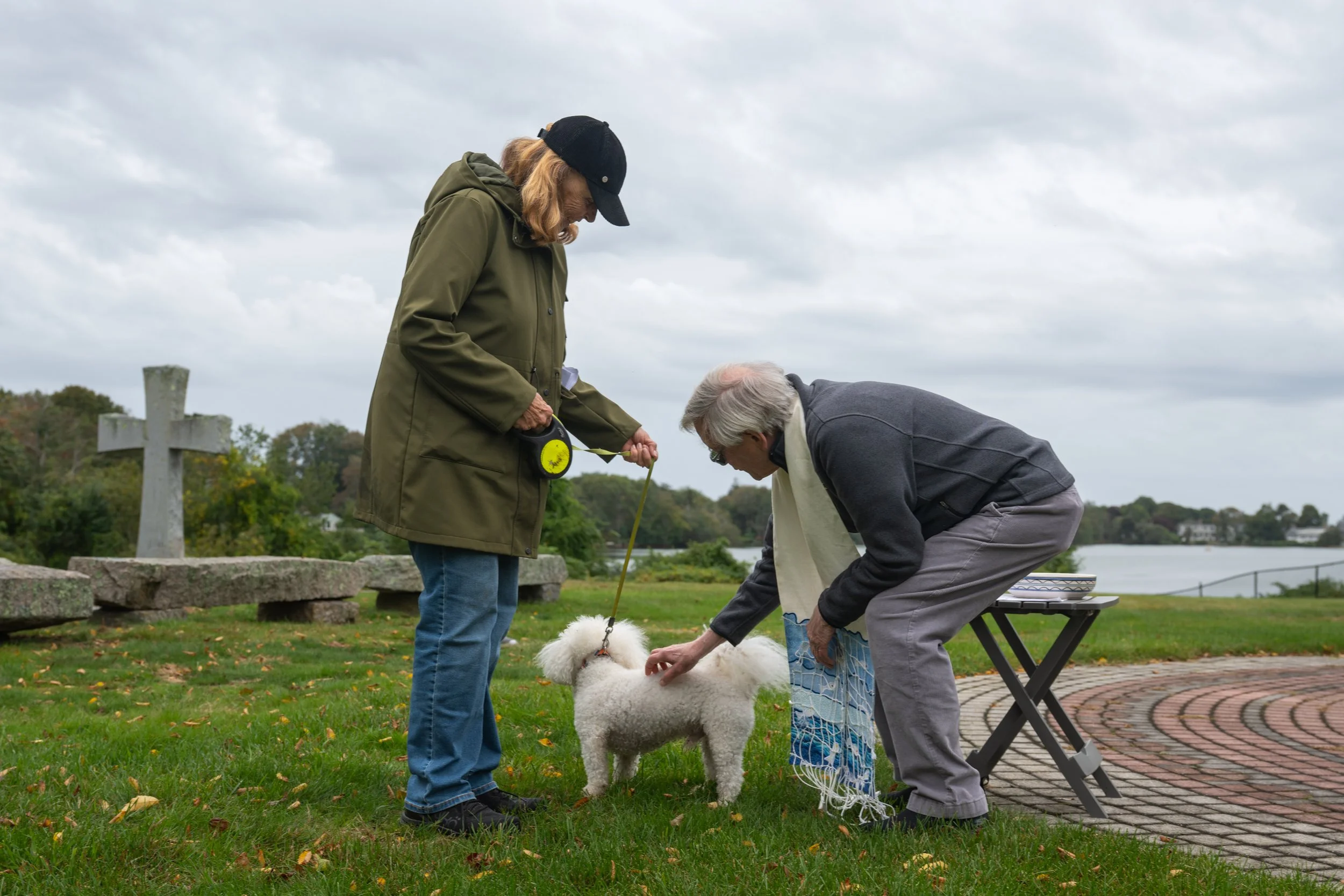 Two older adults with a small white dog outdoors by a lake, with one person holding the leash and the other preparing a dog net.