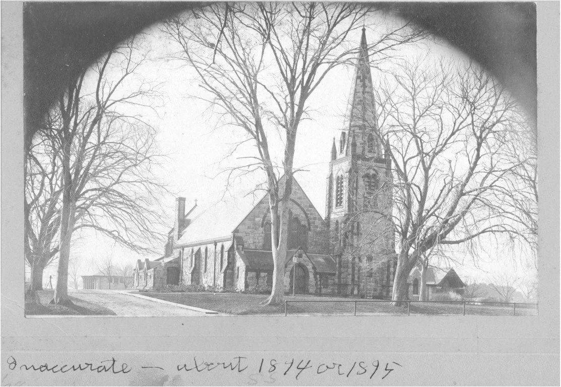 A black and white photograph of St. Barnabas. The church is surrounded by leafless trees and a low fence, with a pathway leading up to it.