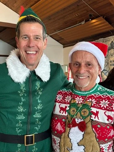 Two smiling men wearing festive Christmas outfits, including a green elf costume and a red holiday sweater with an owl design, posing indoors.