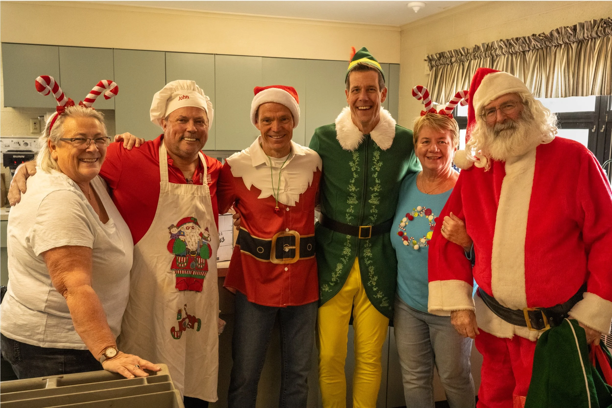 Group of adults wearing festive Christmas costumes, including Santa Claus, an elf, and holiday hats, posing indoors.