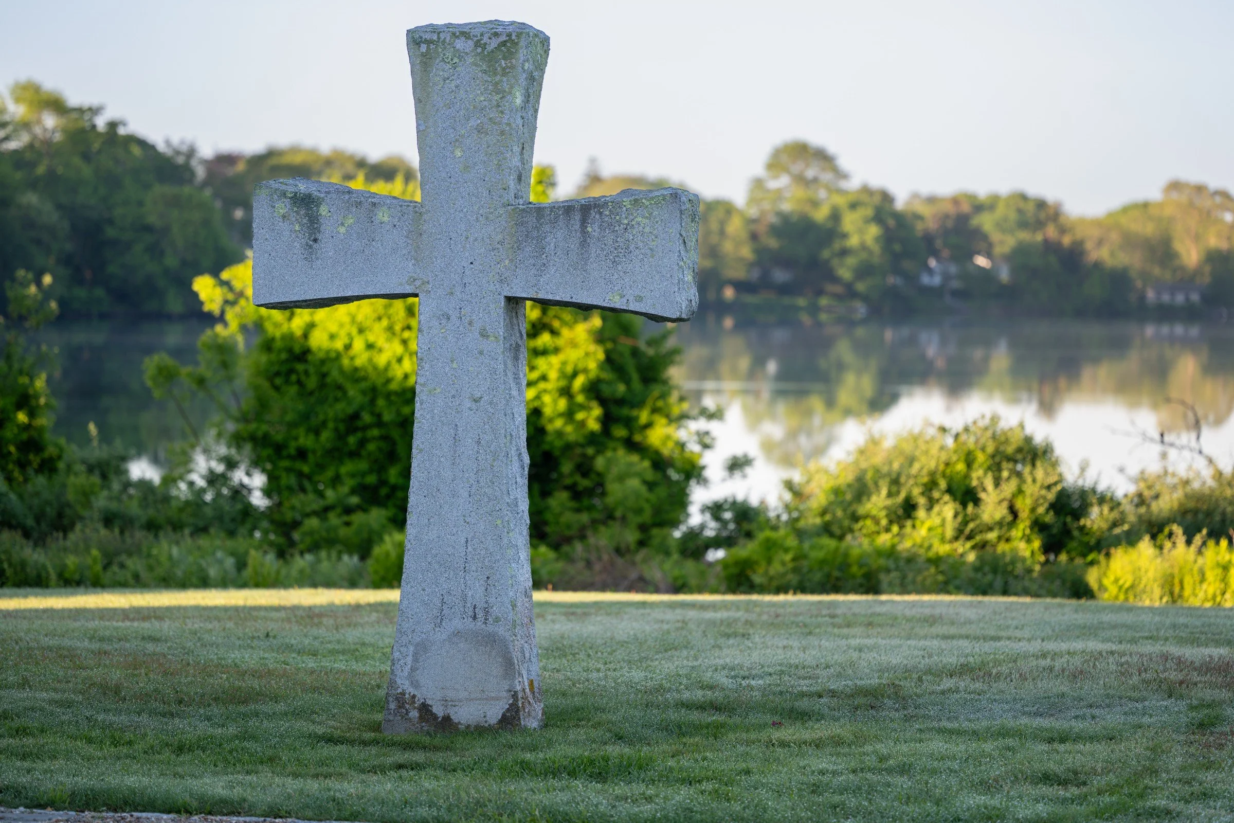 A weathered stone cross monument stands on a grassy area near a water body, with dense green trees and a clear sky in the background.