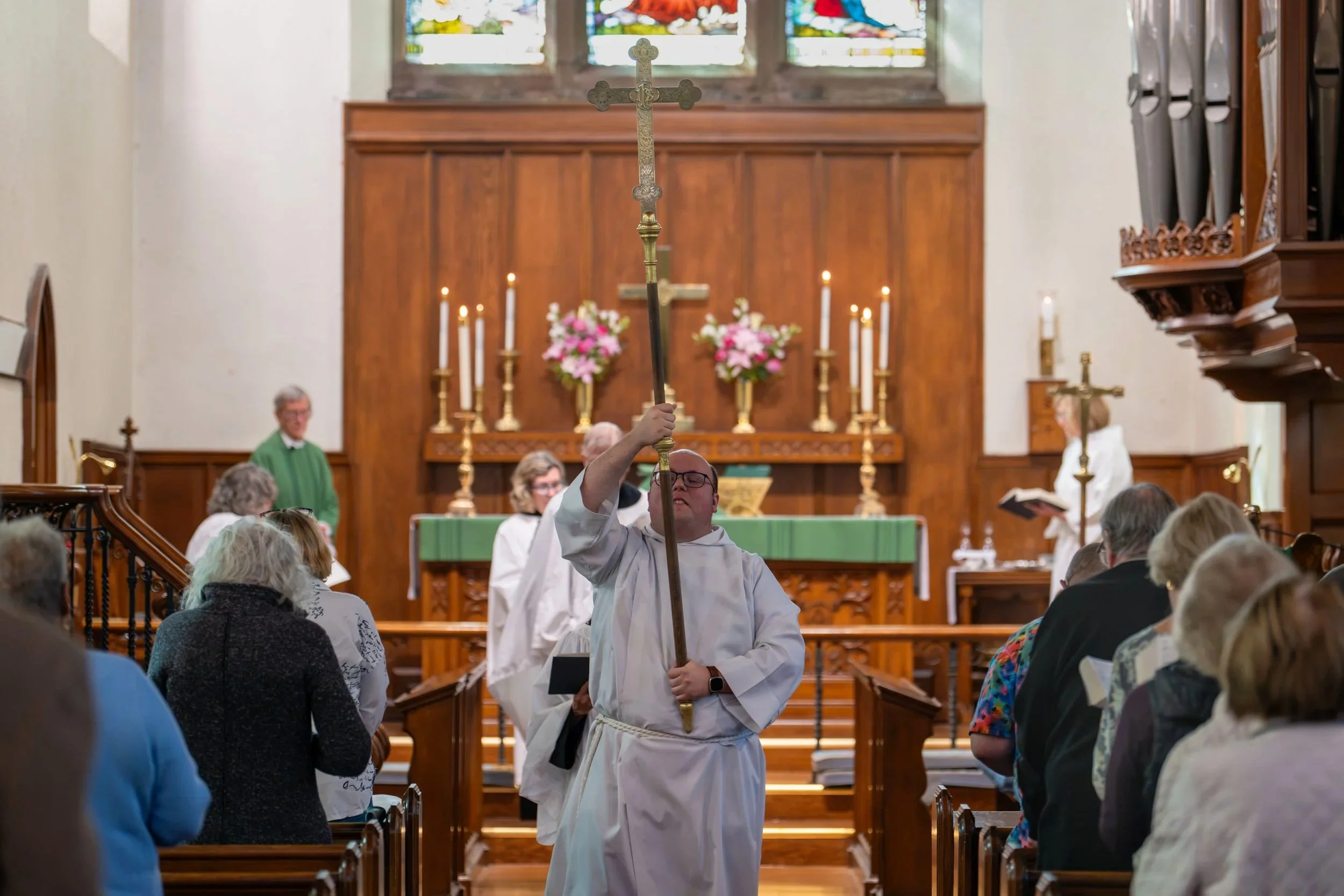 Church procession with a robed acolyte carrying a large cross in the St Barnabas sanctuary, before an altar decorated with candles and flowers.