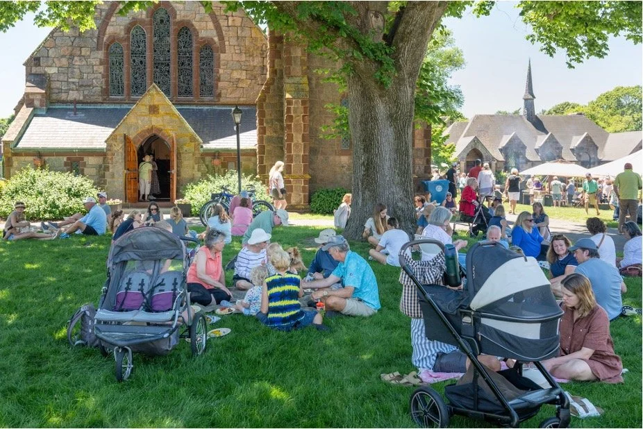Crowd gathered on the lawn outside a stone church at a sunny community festival, with families, children, strollers, and a nearby chapel.