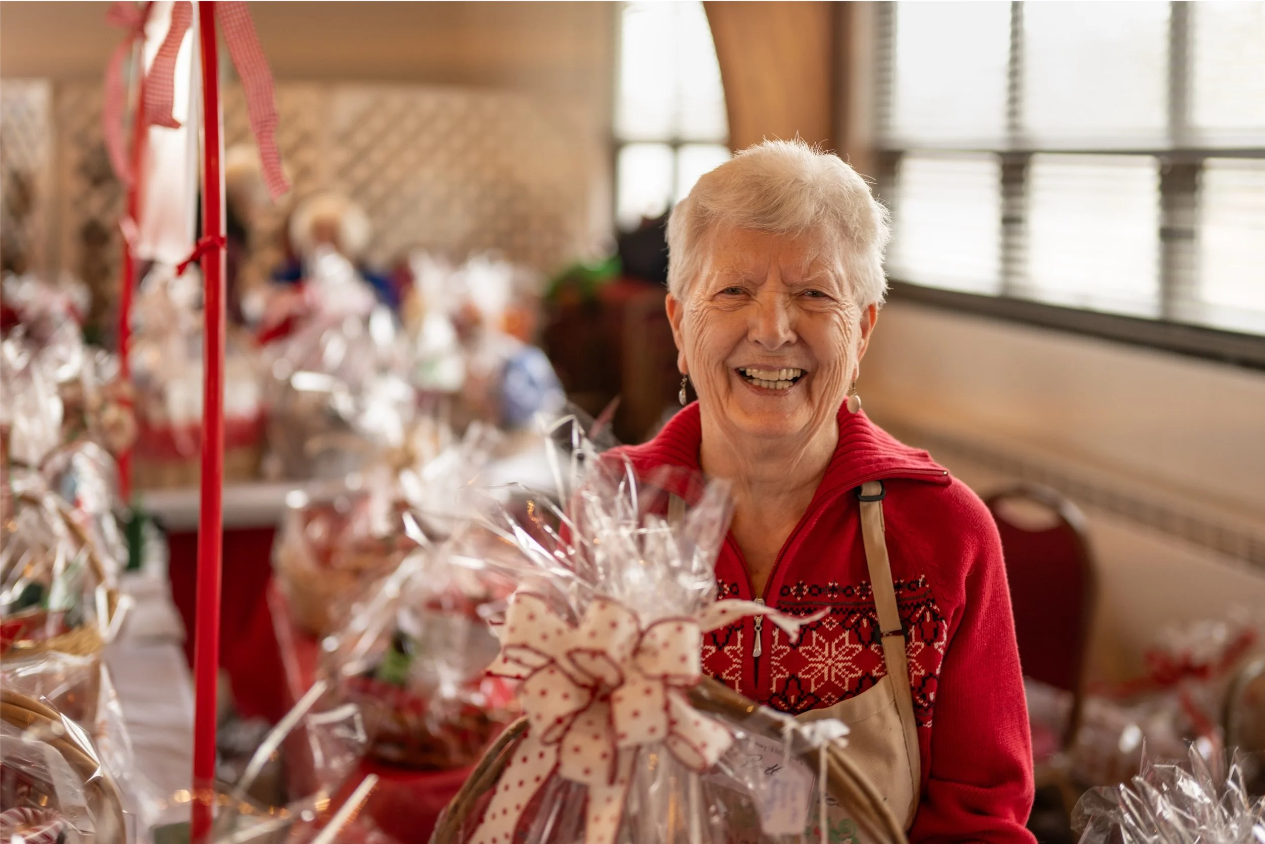 Smiling older woman in a red sweater and apron surrounded by cellophane-wrapped holiday gift baskets.
