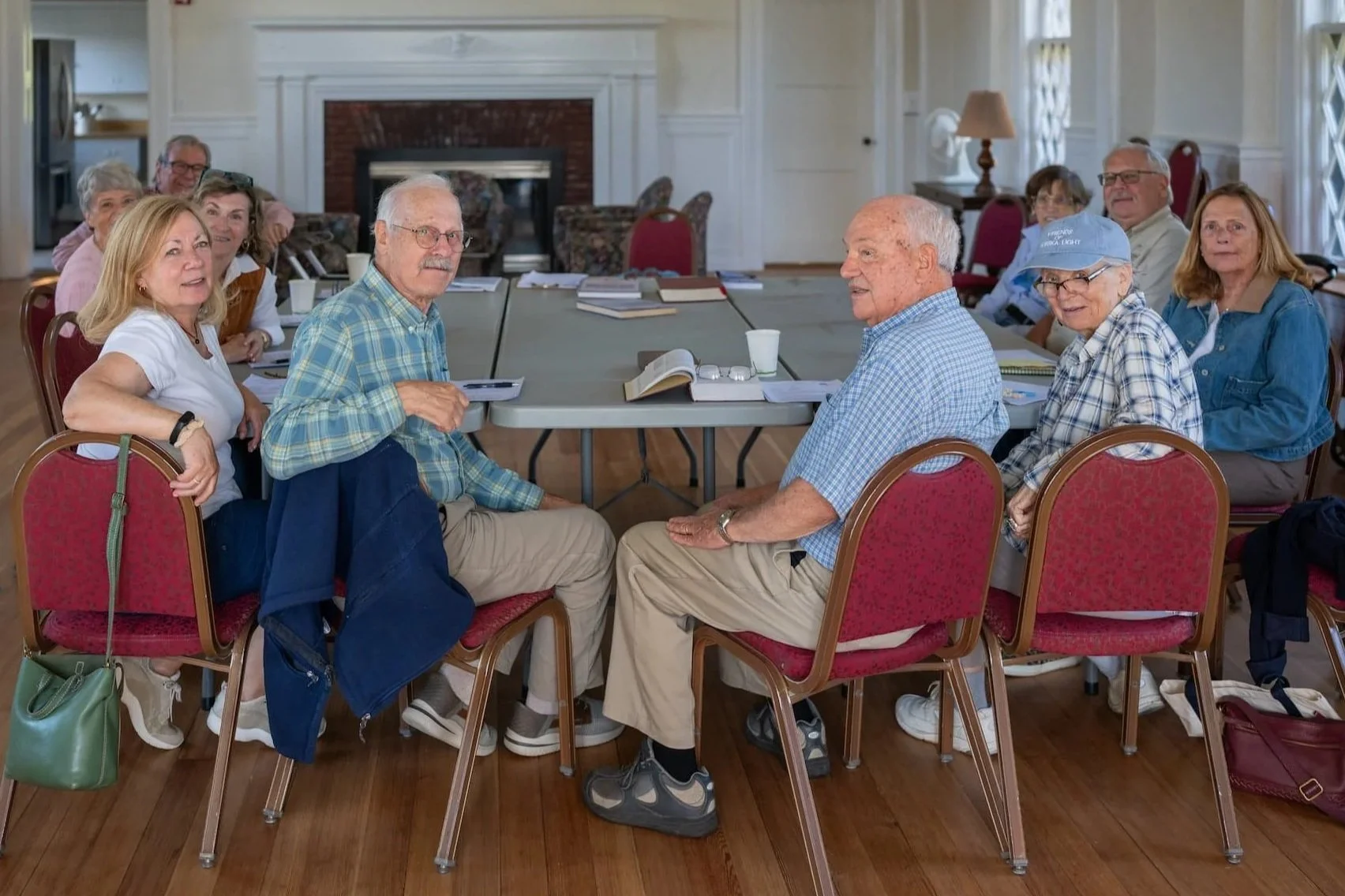A group of parishioners sit around a large rectangular table in a decorated room with white walls, a brick fireplace, and large windows. They are engaged in adult education, with notebooks, books, and cups on the table.