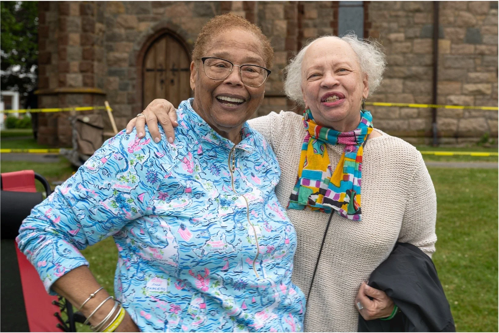 Two smiling older women embracing outdoors in front of a stone church, wearing colorful clothing and scarves.