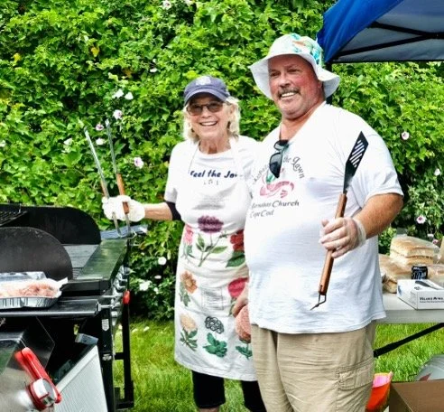 Smiling couple grilling at an outdoor garden barbecue, holding barbecue utensils in front of flowering greenery.