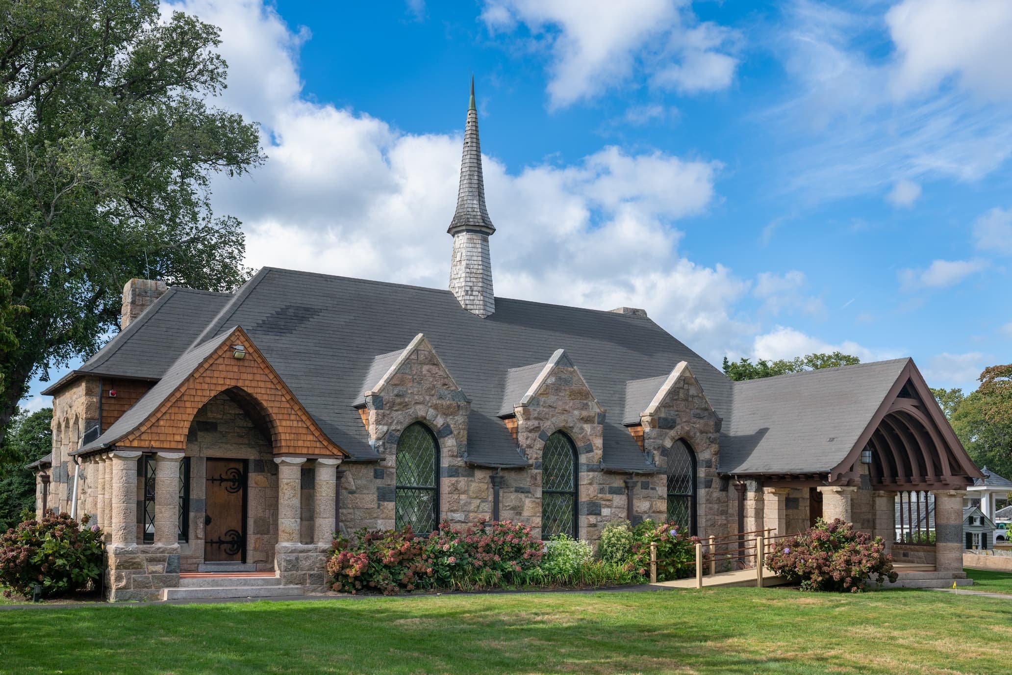 Stone Gothic Revival chapel with a tall shingled steeple, arched stained-glass windows, flower beds, and manicured lawn under a blue sky