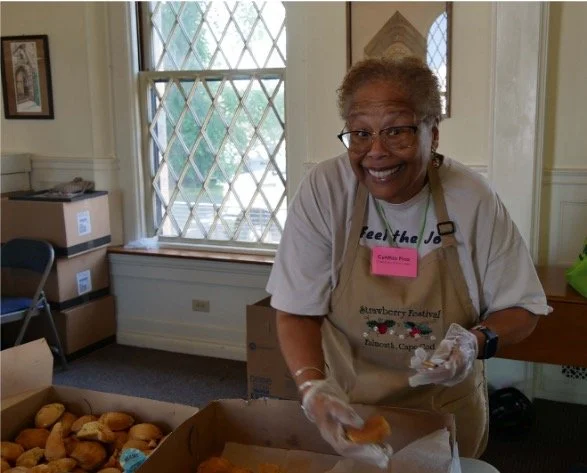 Smiling woman wearing an apron and gloves while handling baked goods at a strawberry festival in a historic building