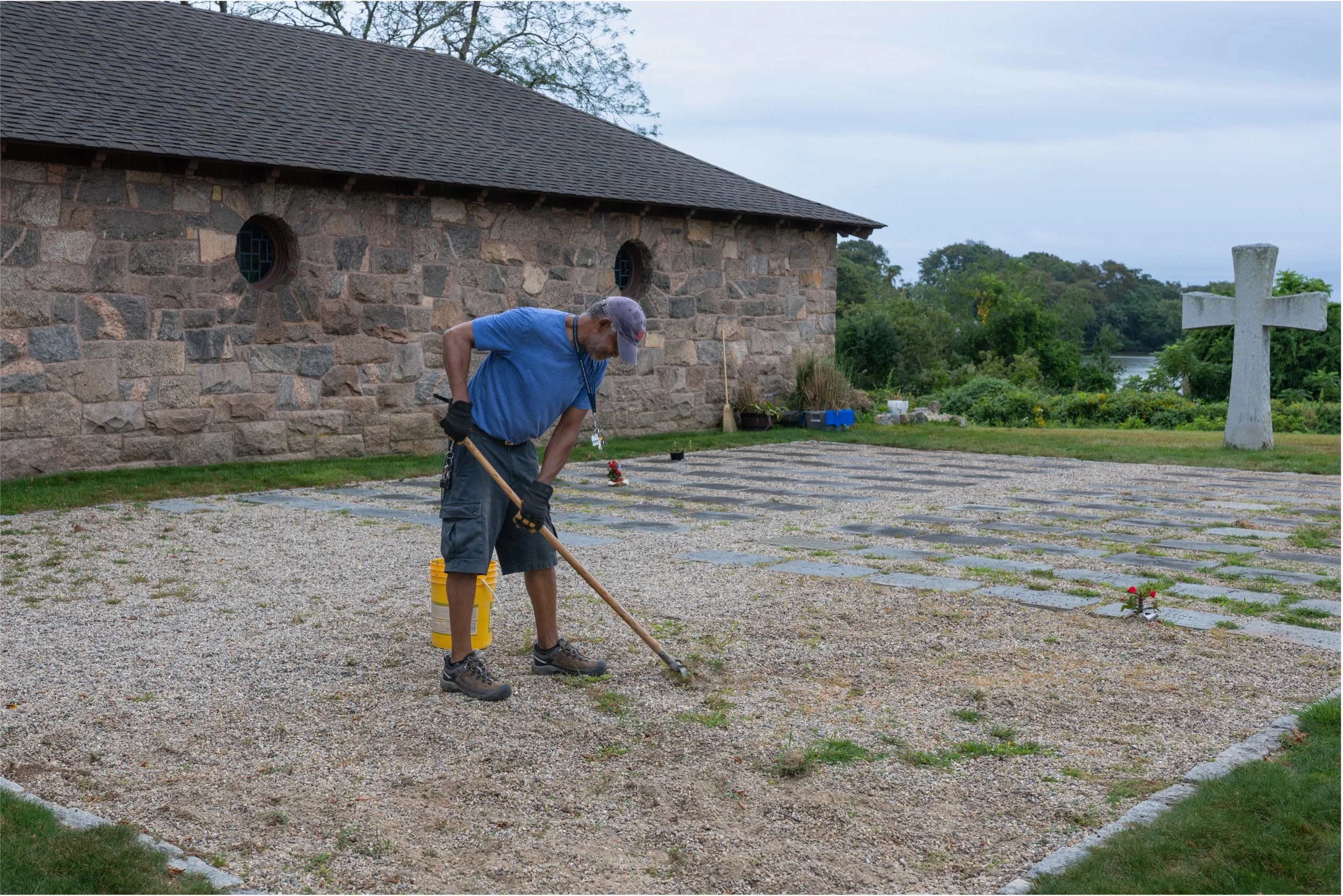 Groundskeeper raking a gravel cemetery beside a historic stone chapel and large white cross