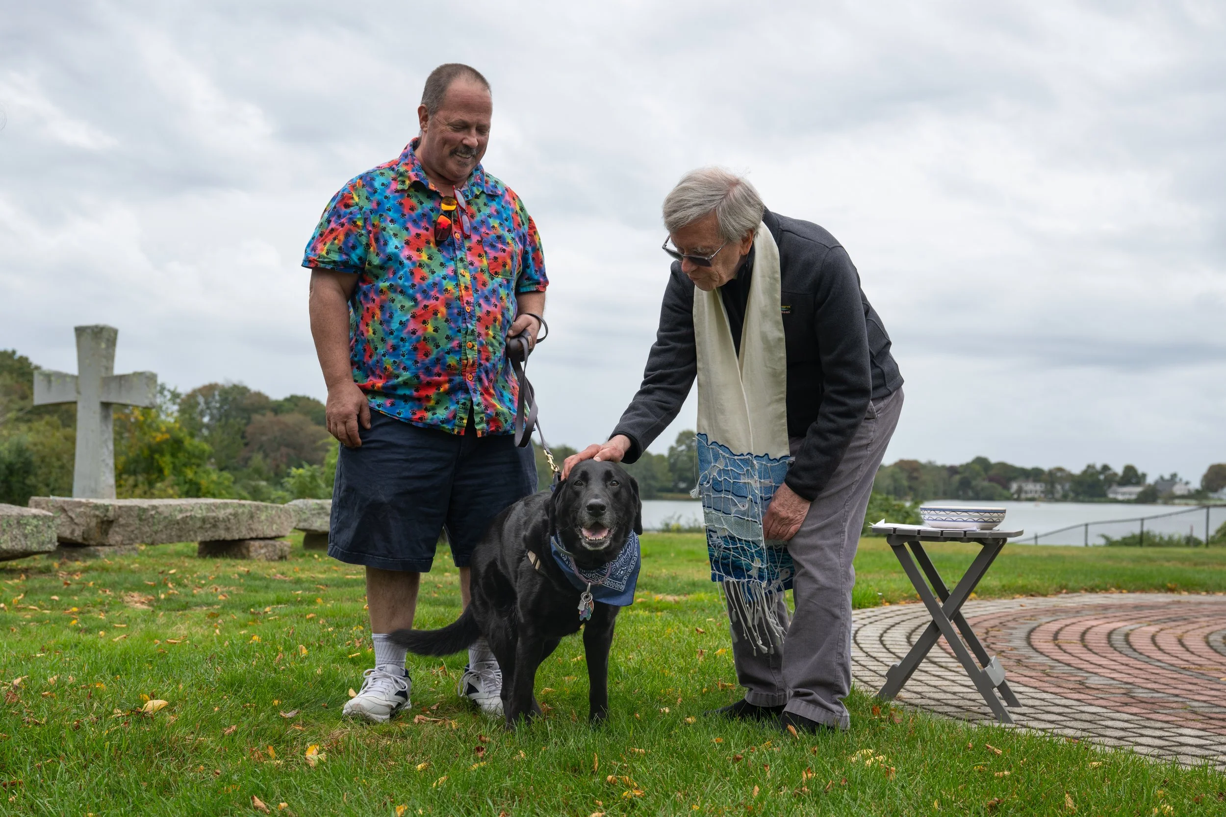Two adults with a black dog at a waterfront park, with a picnic table and stone cross in the background.