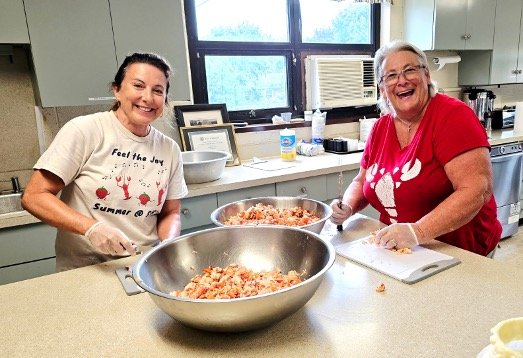 Two women preparing food together in a community kitchen with large mixing bowls and chopped ingredients.