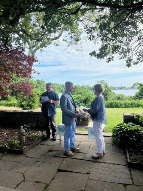 Outdoor wedding ceremony overlooking a lake, with a couple exchanging vows beneath trees and an officiant standing nearby.