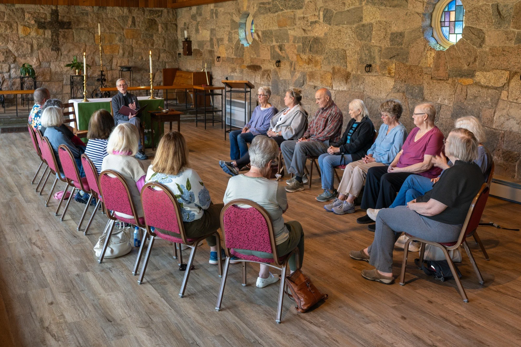 A priest leads a small group of parishioners in prayer in a room with stone walls and small circular stained glass windows, decorated with candles and potted plants.