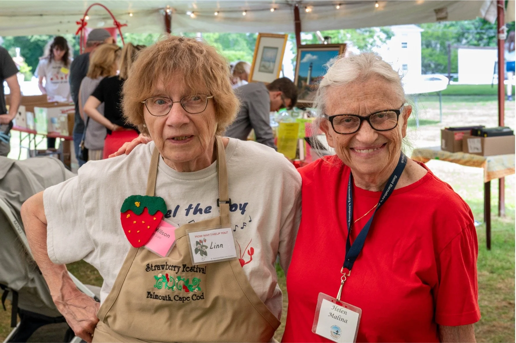 Two smiling older women pose at a strawberry festival booth, with one wearing a festival apron and name badge.