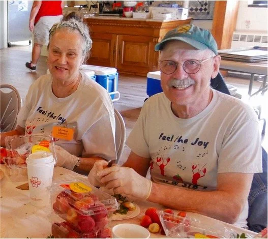 Older couple wearing matching “Feel the Joy” shirts dining together at a table with fruit and drinks.