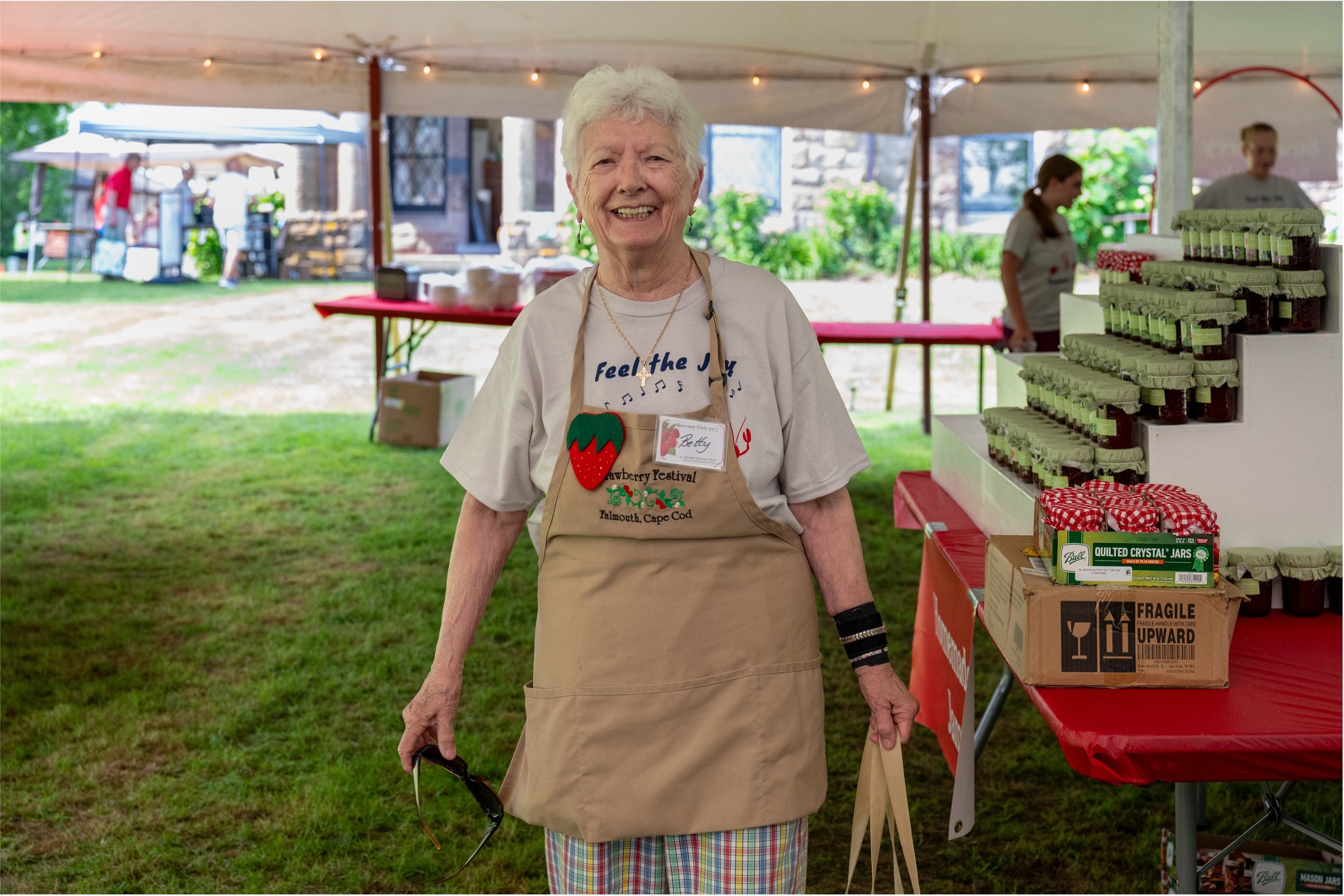 Smiling elderly woman in a strawberry festival apron at a jam and preserves booth under a tent