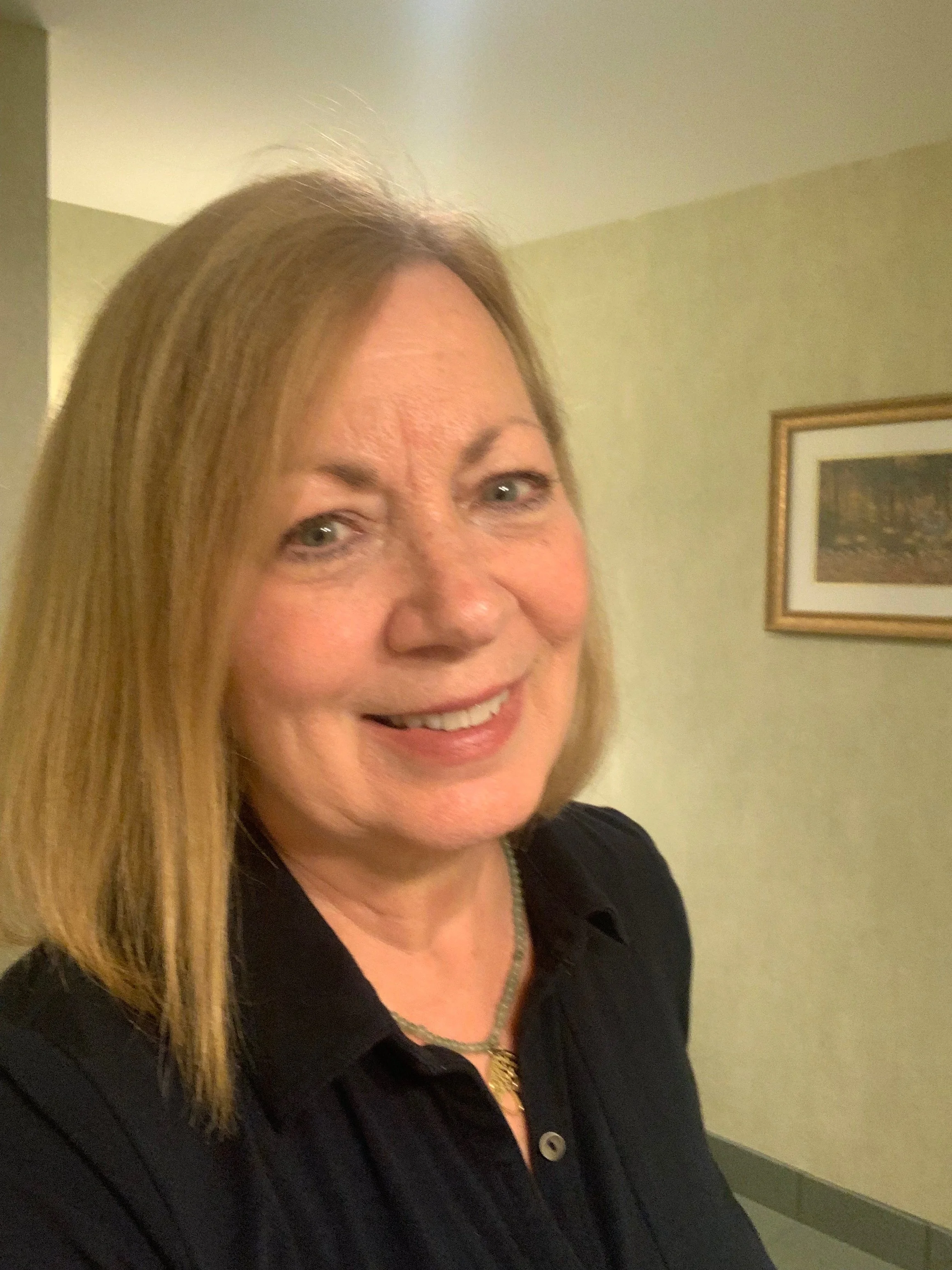 Smiling woman with long blonde hair wearing a black top and necklace indoors