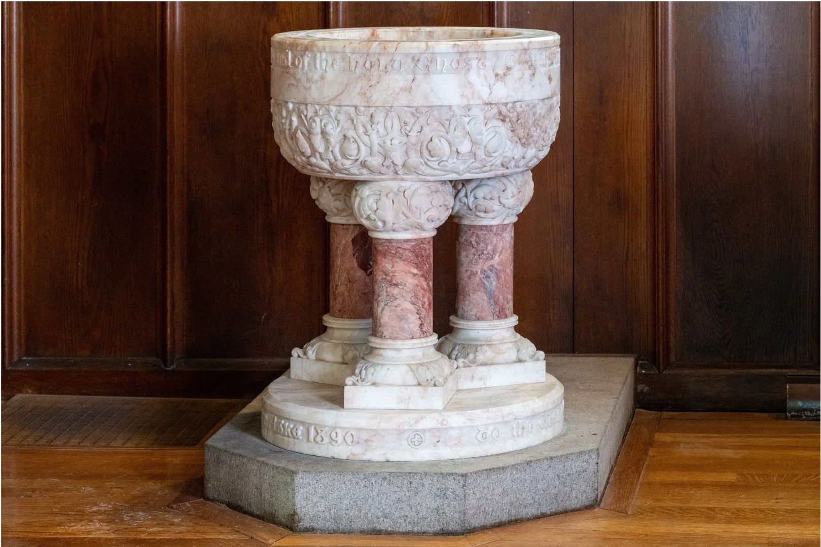 Ornate white marble baptismal font with pink marble columns on a stone pedestal in a wood-paneled church