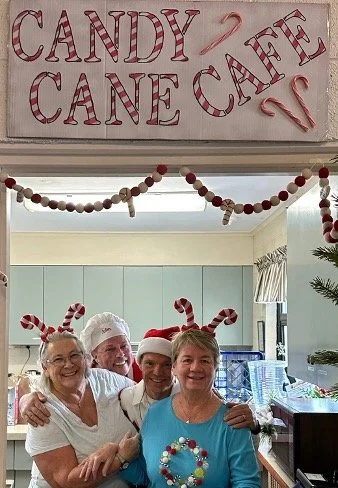 Four smiling people wearing Santa hats and candy cane headbands in a Christmas-decorated kitchen beneath a handmade candy cane cafe sign