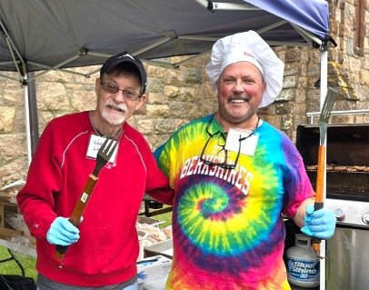 Two smiling men wearing gloves at an outdoor barbecue, with one holding a grill spatula and the other wearing a chef hat beside a grill.