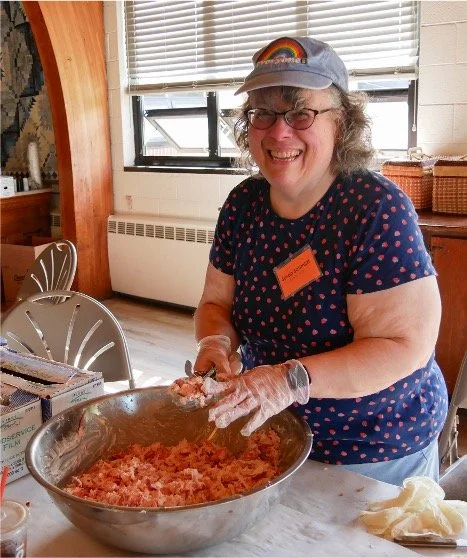 Smiling woman wearing gloves mixes a shredded food mixture in a large stainless steel bowl indoors.