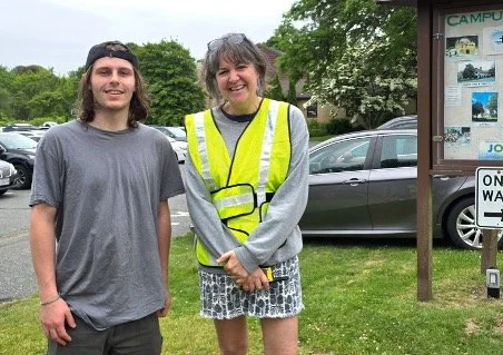 Two people, including a volunteer in a high-visibility vest, pose beside a campus information board in a parking lot.