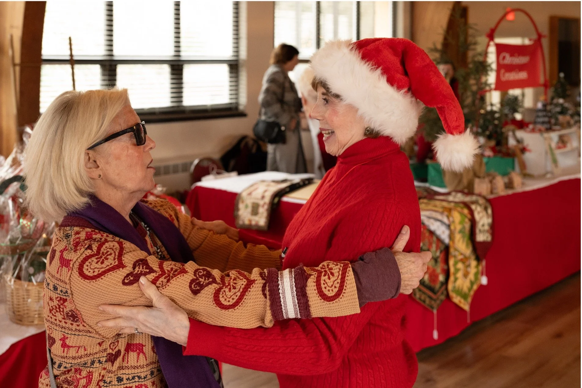 Older women embracing at a Christmas celebration, with one wearing a Santa hat and a decorated Christmas tree in the background.