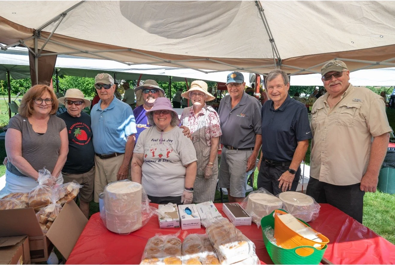 Group of volunteers posing at a community event booth with baked goods and refreshments under a canopy.