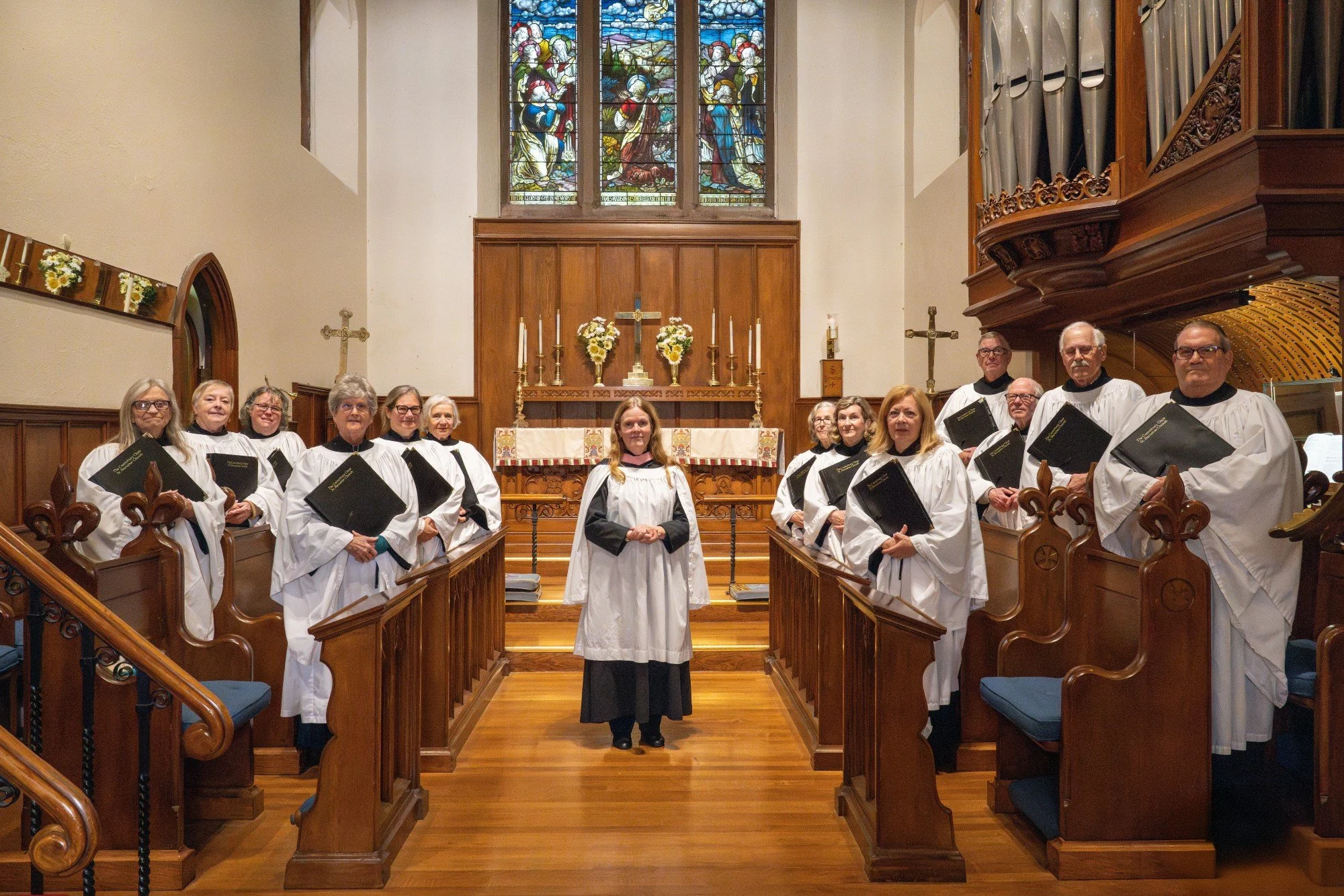 A church choir standing inside the church, dressed in white robes with black collars, holding hymn books,smiling. The church interior features wooden pews, a stained glass window depicting religious figures, and an altar adorned with flowers, candles, and religious symbols.