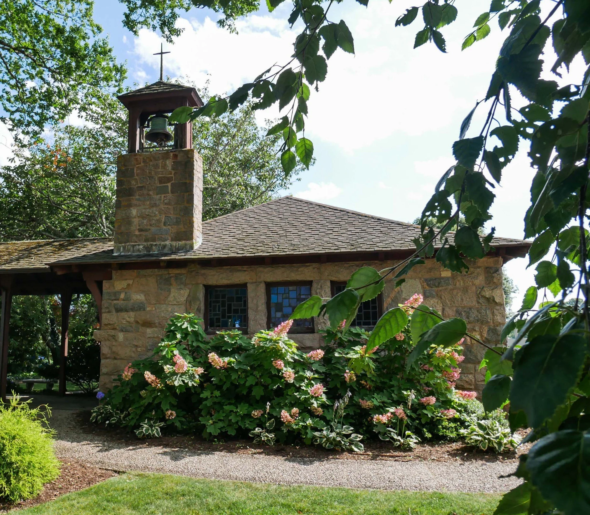 Stone chapel with a bell tower and cross surrounded by flowering shrubs and leafy trees