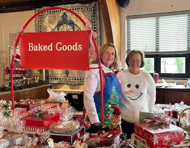 Two women stand behind a festive holiday table filled with packaged baked goods beneath a red “Baked Goods” sign.