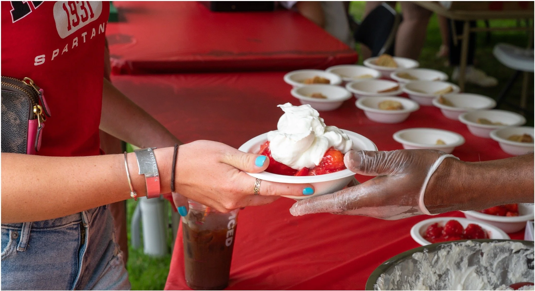 Vendor in gloves hands a bowl of strawberries topped with whipped cream at an outdoor dessert stand