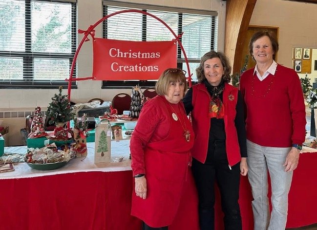 Three women pose at a Christmas Creations craft sale table decorated with holiday gifts and treats.